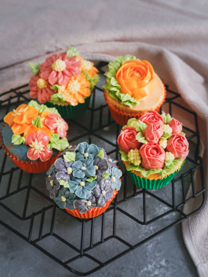 Five decorated cupcakes with flowers made from buttercream on a black wire rack